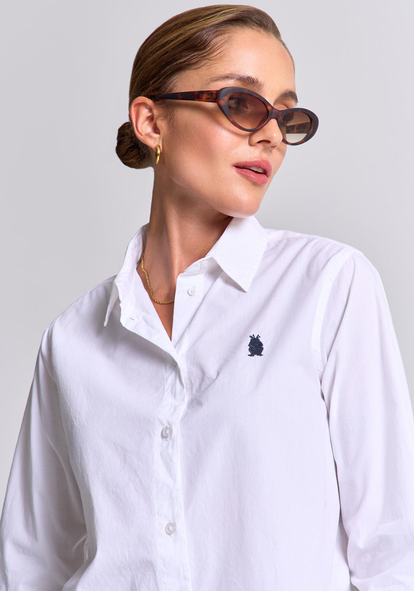 A woman with slicked-back hair wears dark oval sunglasses, gold hoop earrings, and a white Princess Goes Hollywood Regular Fit Baumwollhemd mit Polokragen, featuring a small black logo on the chest, against a light gray background.
