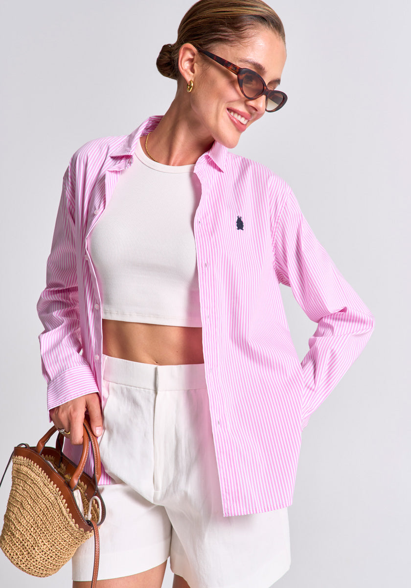 A woman in a Princess Goes Hollywood striped cotton shirt, worn regular fit over a white crop top with high-waisted shorts, smiles to the side. She accessorizes with sunglasses, a woven handbag, and a sleek bun against a plain light background.