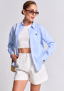 A woman poses confidently in Princess Goes Hollywood cat-eye sunglasses, a blue-striped Regular Fit Baumwollhemd with Polokragen over a white crop top, high-waisted white shorts, and carries a straw bag. Her hair is tied back against a plain light gray background.