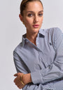 A woman with slicked-back hair wears a gestreiftes Baumwollhemd in Regular Fit mit Polokragen von Princess Goes Hollywood, goldenen Creolen und einem dünnen Armreif. Sie blickt selbstbewusst vor grauem Hintergrund, die Arme locker verschränkt.