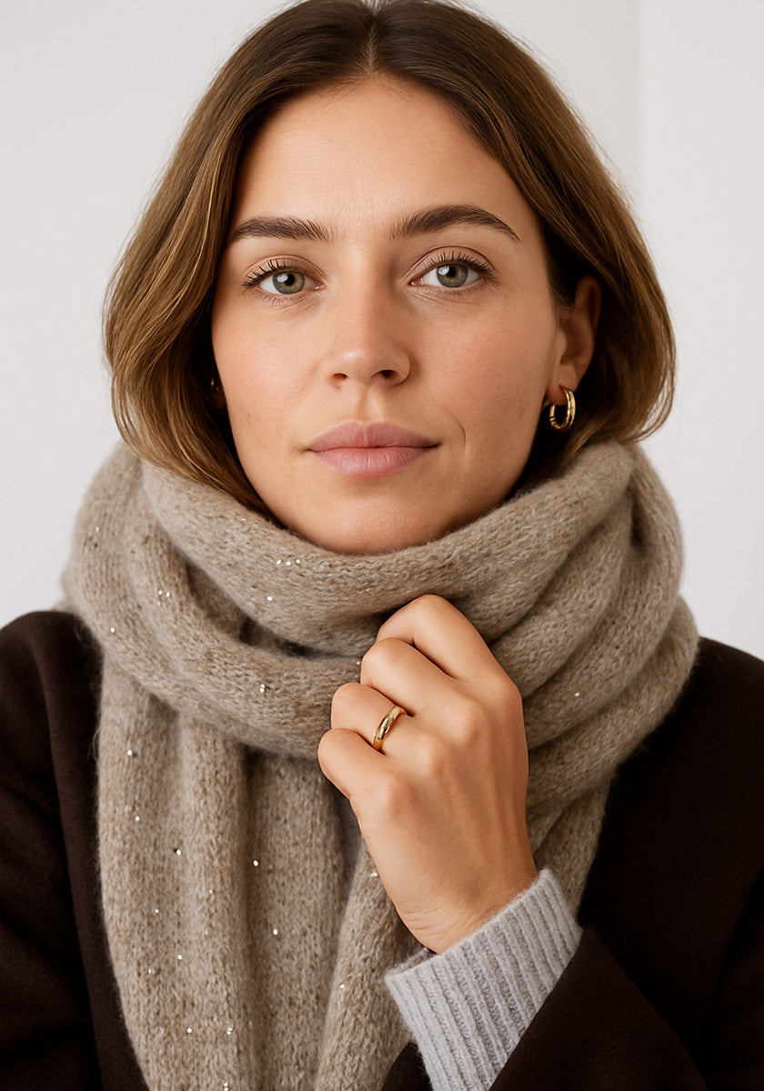 A woman with light brown hair and green eyes gazes at the camera. She wears gold jewelry, a dark coat, and gently holds a sparkling Princess Goes Hollywood Merino-Kaschmir-Schal with subtle Strassverzierung.