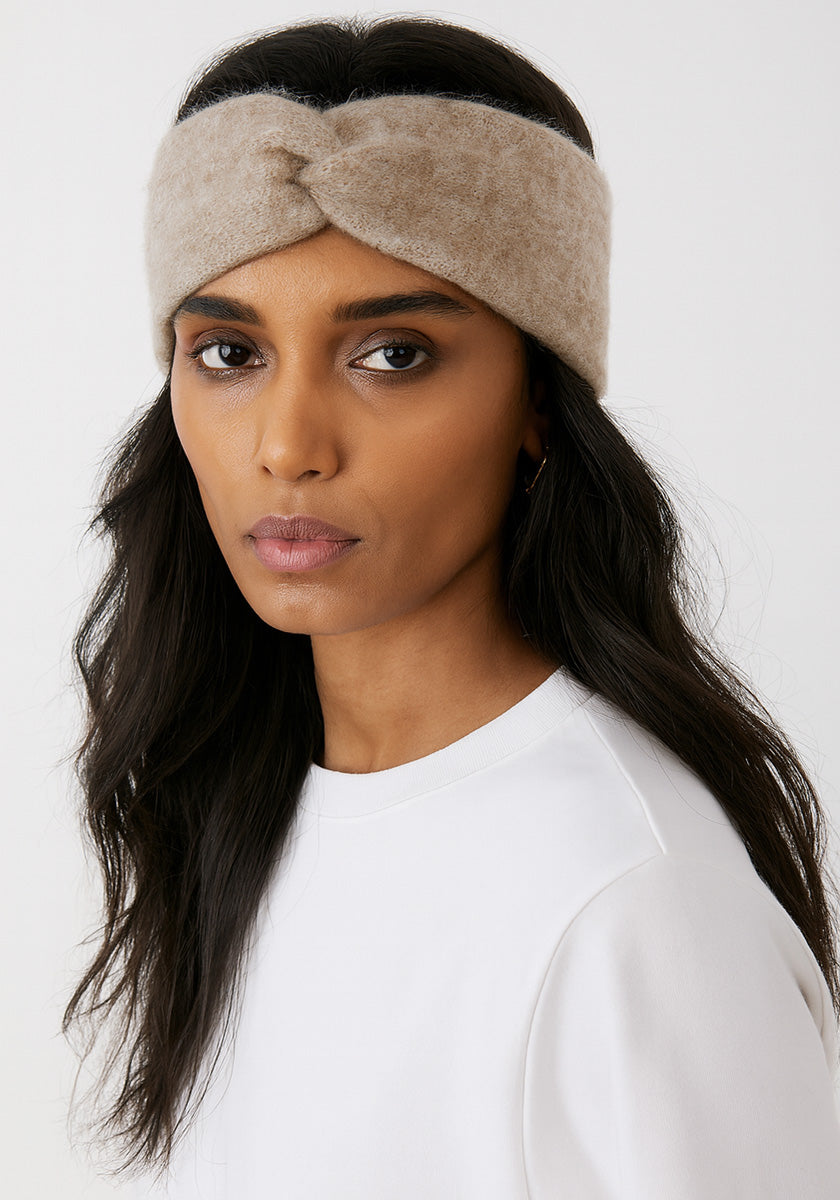 A woman with long, dark wavy hair wears a light beige, fuzzy Kaschmir Stirnband with twist detail from Princess Goes Hollywood. Dressed in a plain white top, she faces forward with a neutral expression against a white background.