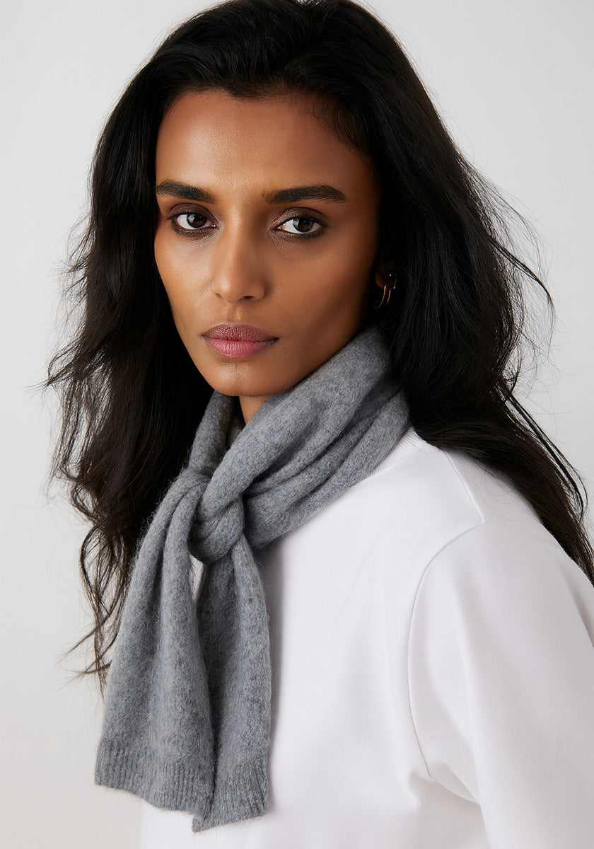 A woman with long, dark hair and medium skin tone wears a Princess Goes Hollywood 100% Kaschmir Schal loosely around her neck over a white top, looking at the camera with a neutral expression against a plain, light background.