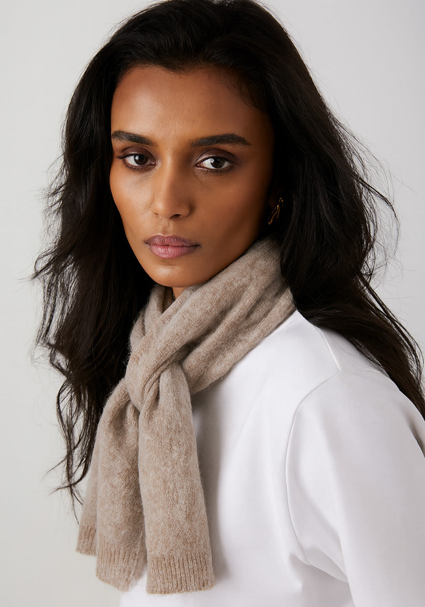 A woman with long dark hair and brown skin looks intently at the camera, wearing a beige Princess Goes Hollywood Kaschmir Schal draped over a white top. The plain, light background accentuates her serious expression.