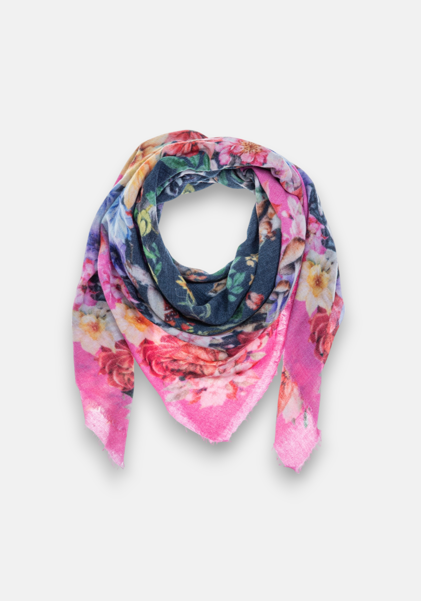 The Princess Goes Hollywood triangular scarf features vibrant pink, yellow, and green floral patterns on a dark blue background, styled in a loose fold on a light grey surface. This lightweight scarf has frayed edges and an exceptionally soft feel.