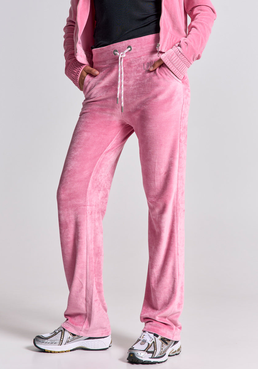 A person stands with hands in pockets, wearing Princess Goes Hollywood light pink Nicki-Stoff track pants with a drawstring waist and matching jacket, paired with white and gray sneakers. The plain background highlights the stylish outfit; head not visible.
