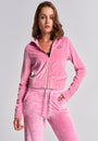 A woman with shoulder-length brown hair wears a light pink Princess Goes Hollywood velour zip-up hoodie and matching sweatpants, facing forward with one hand in her pocket against a gray background, giving a confident, relaxed expression.