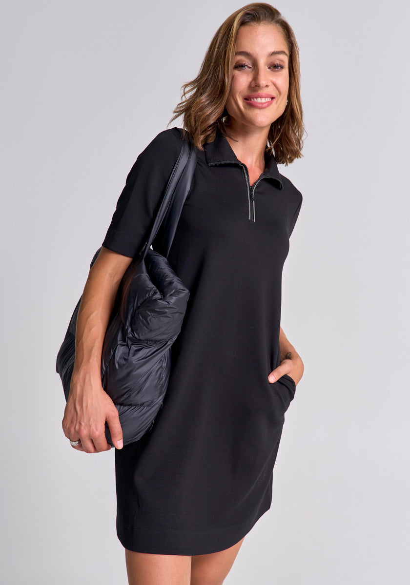 A woman with light brown hair wears a black Mini Sweatkleid with Reverskragen by Princess Goes Hollywood and carries a large, puffy black shoulder bag. She stands against a light gray background, smiling with one hand in her pocket.