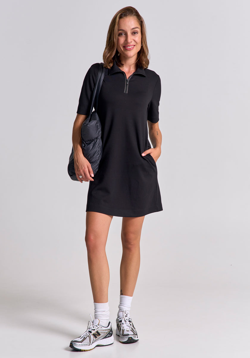 A woman with shoulder-length brown hair smiles against a plain background, wearing a Princess Goes Hollywood short-sleeved Mini Sweatkleid with front zipper, white socks, athletic sneakers, and carrying a black shoulder bag. One hand is in her pocket.