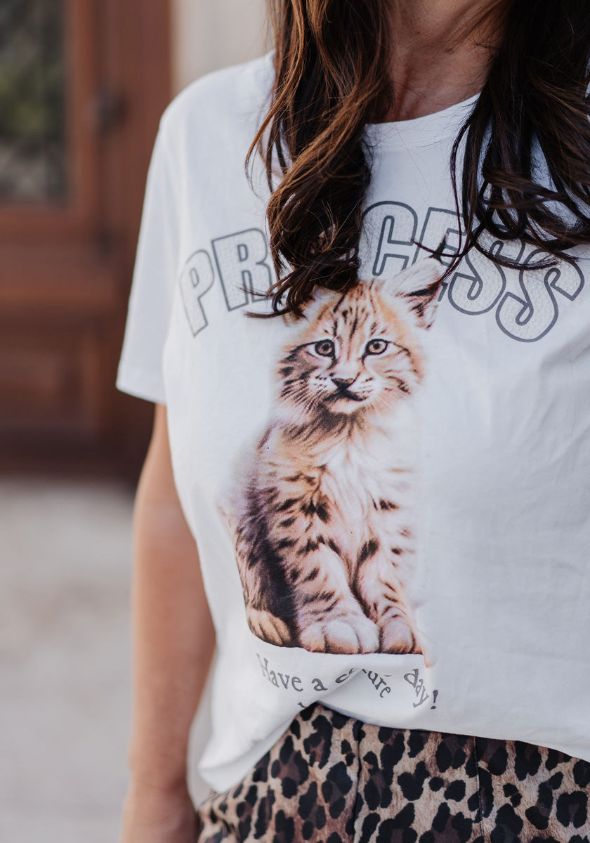 A woman with long brown hair wears a Princess Goes Hollywood white cotton crew-neck T-shirt featuring a tabby kitten photo and “PRINCESS,” styled with a lynx-print skirt; her face is partially out of frame.