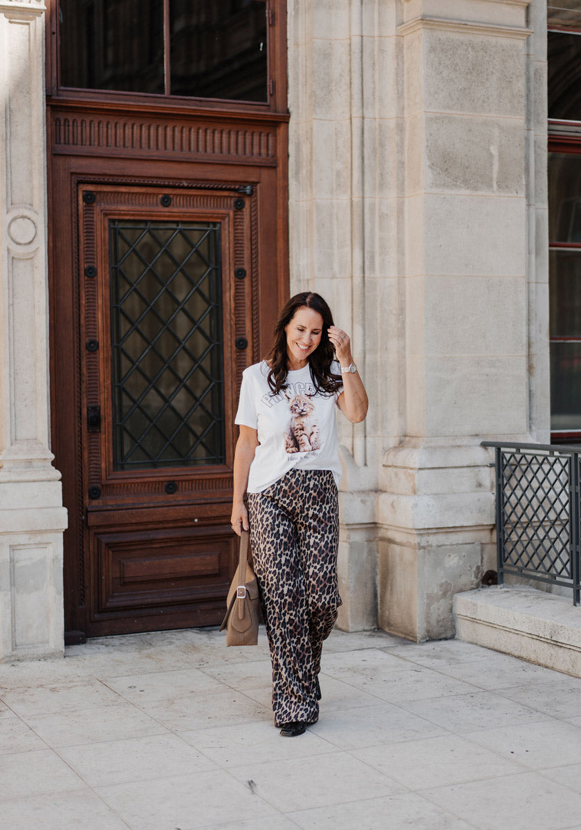 A woman with long dark hair walks outside near a stone building, wearing a Princess Goes Hollywood weißes Baumwoll T-Shirt mit Rundhalsausschnitt and loose Luchs-Print pants. She carries a tan handbag and smiles as she touches her hair.