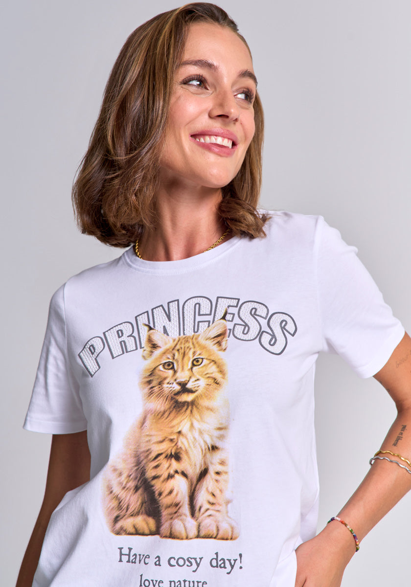 A smiling woman with shoulder-length brown hair models a Princess Goes Hollywood Baumwolle T-Shirt featuring a Luchs-Print, “PRINCESS” text above, and “Have a cosy day! love nature.” below. The Rundhalsausschnitt offers a classic look. Light gray background.
