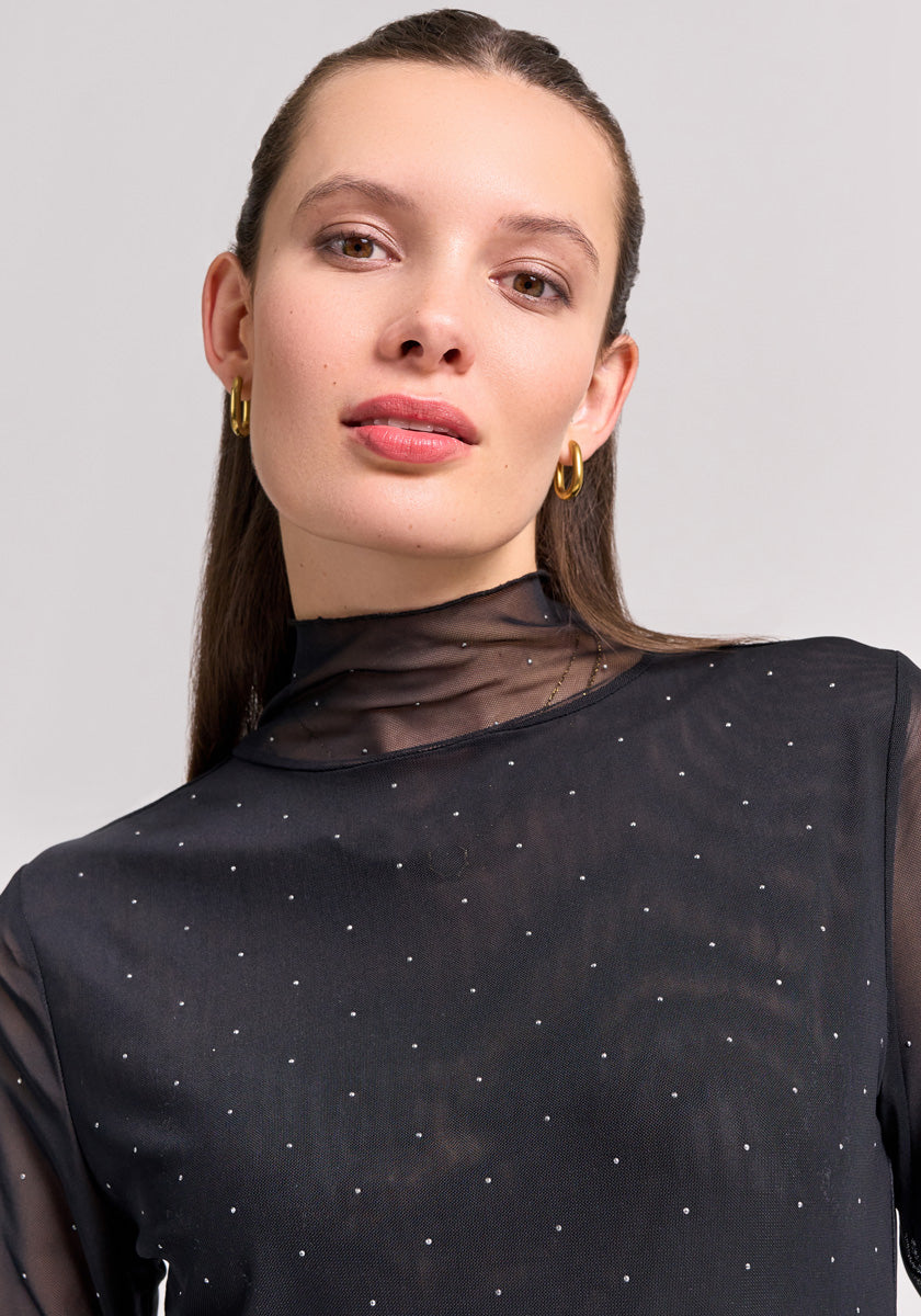 A woman with straight brown hair, gold hoop earrings, and natural makeup models a black mesh shirt with small dots and a stand-up collar by Princess Goes Hollywood. She faces the camera neutrally against a plain light gray background.