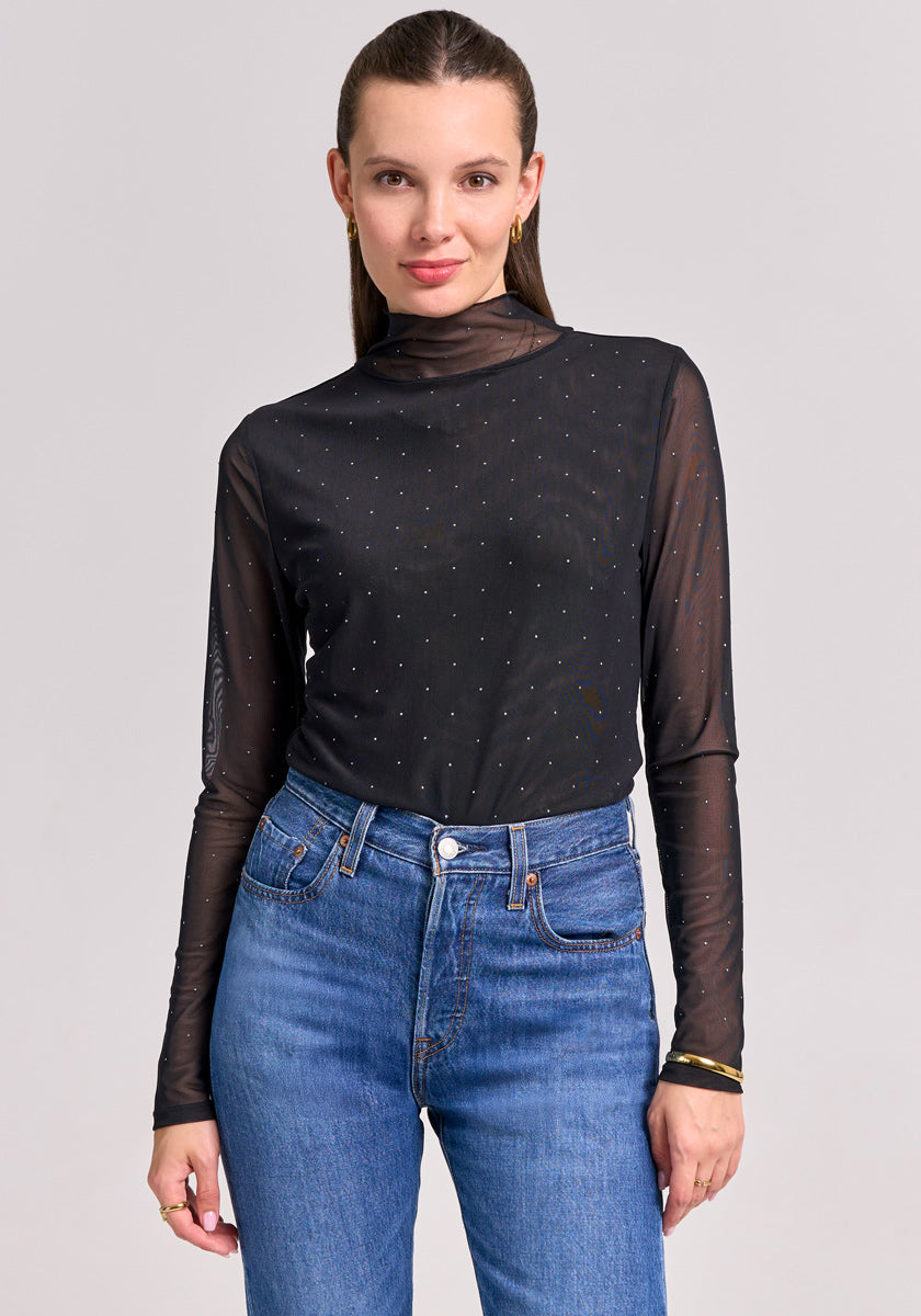A woman faces forward in a sheer Princess Goes Hollywood Mesh-Shirt with subtle rhinestone details and high-waisted blue jeans. Her straight, light brown hair is pulled back; she wears minimal makeup and stands before a plain, light background.