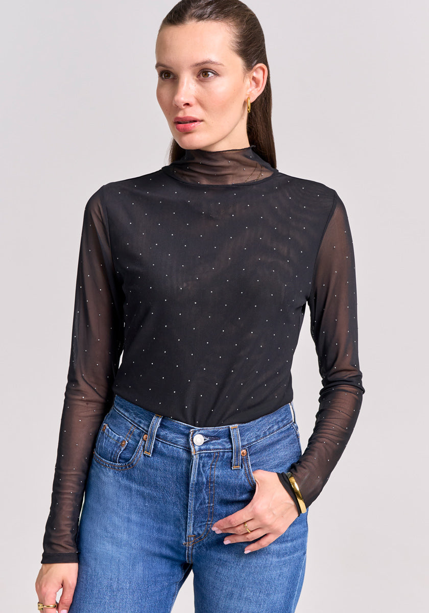A woman with straight brown hair in a low ponytail wears a sheer schwarzes Mesh-Shirt mit Stehkragen und weißen Punkten von Princess Goes Hollywood, kombiniert mit blauer High-Waist-Jeans, vor grauem Hintergrund mit neutralem Blick.