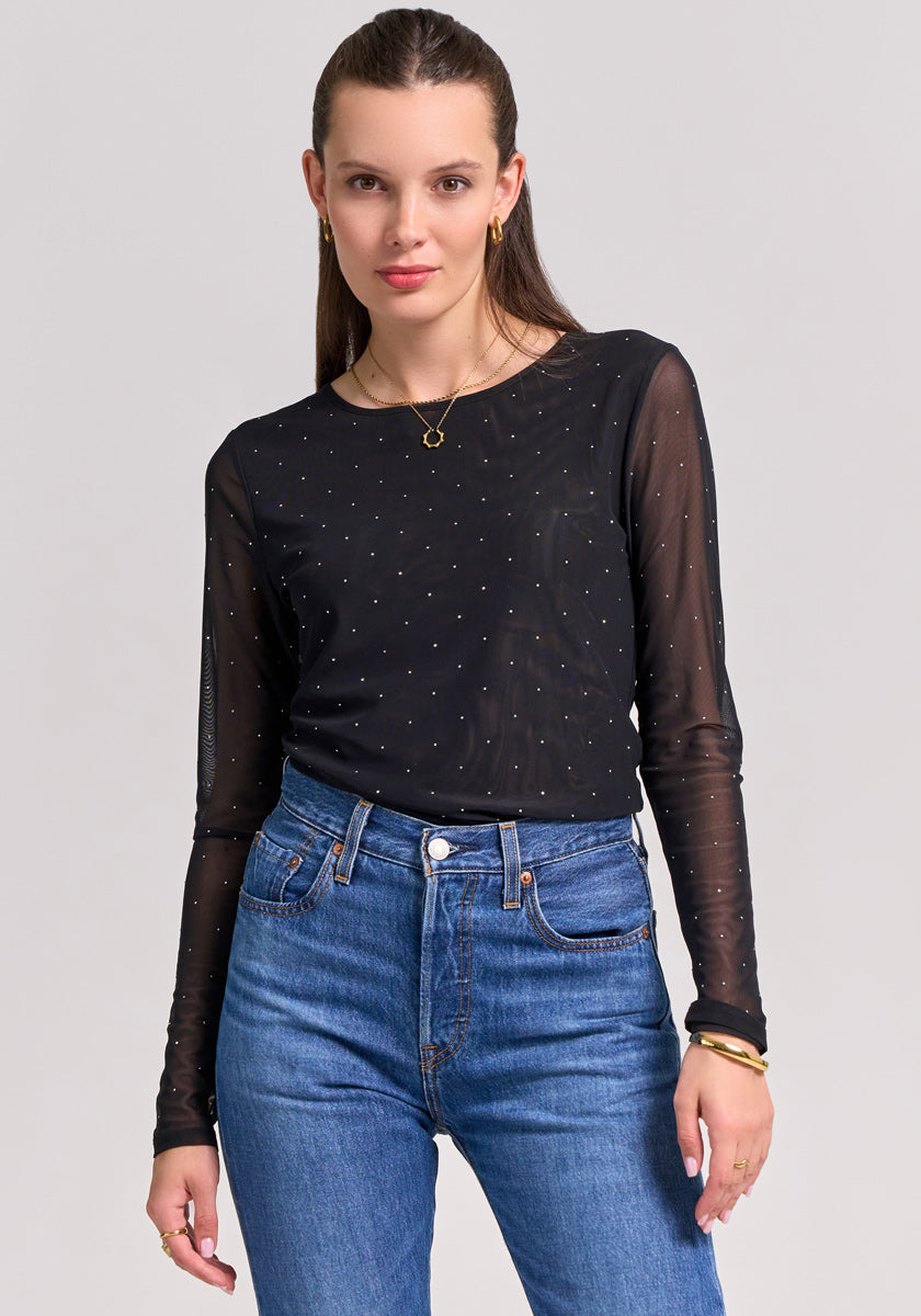 A woman with straight brown hair wears a Princess Goes Hollywood Mesh-Shirt with transparent sleeves and dotted patterns, tucked into high-waisted blue jeans. She accessorizes with gold hoop earrings, a necklace, and bracelets against a light background.
