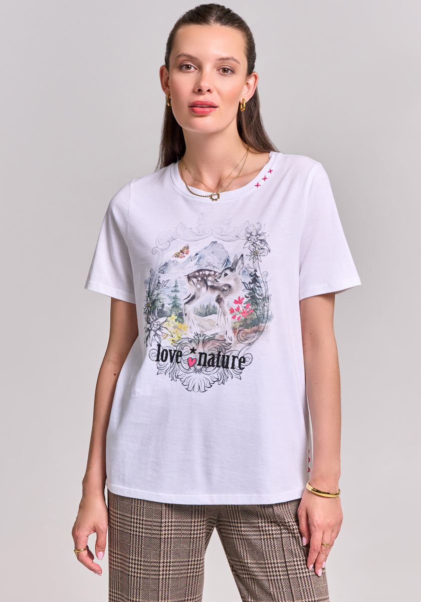 A woman with straight brown hair wears gold earrings, a necklace, plaid pants, and a white Princess Goes Hollywood Baumwoll T-Shirt featuring a printed wolf, flowers, mountains, and love nature, standing against a light gray background.