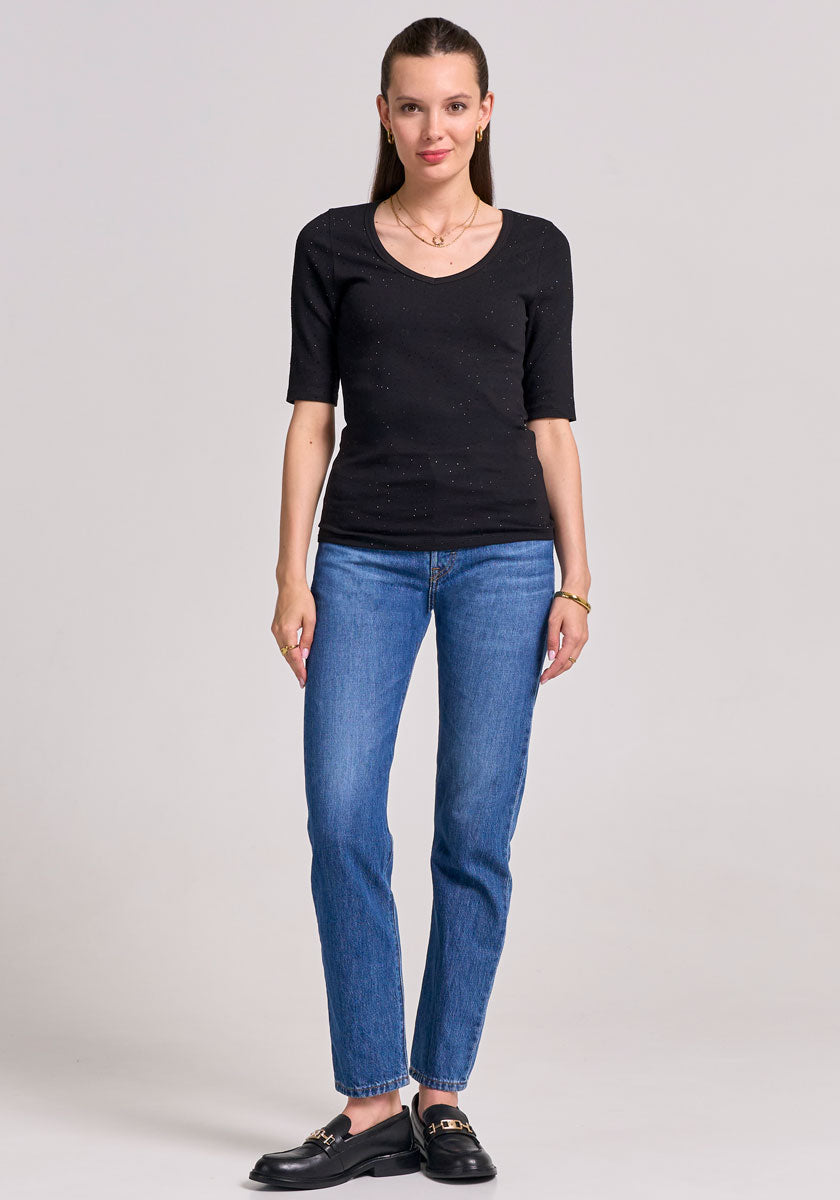 A woman with straight brown hair tied back stands against a light gray background, wearing a Princess Goes Hollywood black V-Ausschnitt Baumwoll-Shirt, blue straight-leg jeans, black loafers, gold necklaces, small earrings, and a wristwatch.
