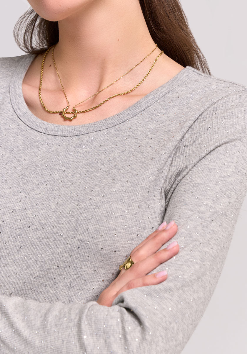 A woman with long brown hair wears a light grey Princess Goes Hollywood Baumwoll-Shirt with Stretchanteil and subtle Strassverzierung. She stands with arms crossed, two gold necklaces, and a gold ring, her face partially visible.