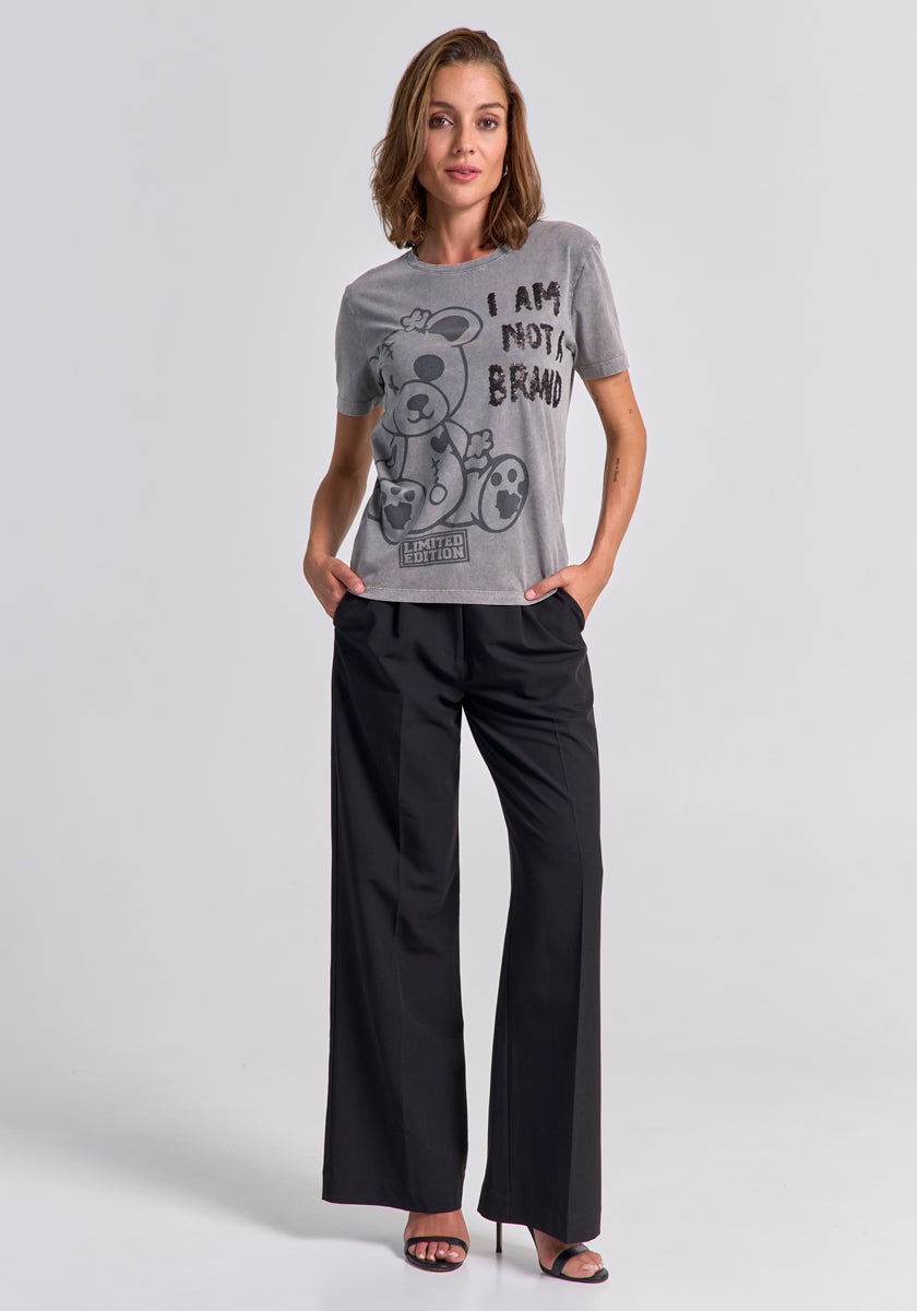 A woman stands in a studio wearing a gray Princess Goes Hollywood Statement Shirt with a bear graphic and "I AM NOT A BRAND" text, paired with black wide-leg trousers and heeled sandals. Her look is relaxed and confident.