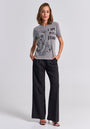 A woman stands in a studio wearing a gray Princess Goes Hollywood Statement Shirt with a bear graphic and "I AM NOT A BRAND" text, paired with black wide-leg trousers and heeled sandals. Her look is relaxed and confident.