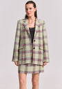 A woman faces forward in a cream, black, pink, and green Karo-Print Tweed Blazer and skirt by Princess Goes Hollywood. She pairs them with a black top, minimal makeup, pulled-back hair, and a simple gold necklace on a light gray background.