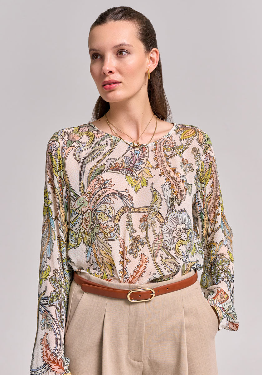 A woman with long brown hair slicked back wears a Princess Goes Hollywood loose blouse featuring a green, orange, and beige floral Paisley print. She pairs it with tan high-waisted pants and a brown belt, gazing calmly to the side.