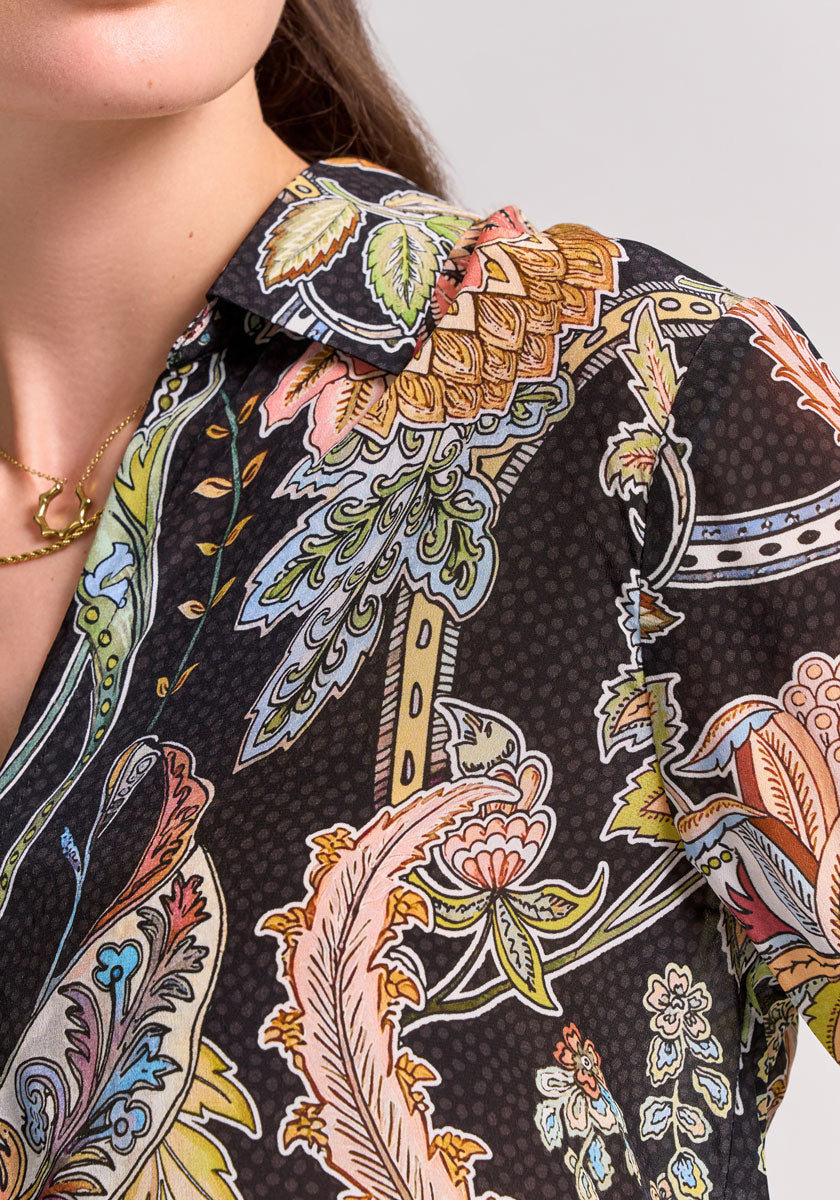 A close-up shows a person wearing a Princess Goes Hollywood Damenbluse with vibrant floral and paisley prints on black. Part of the face, brown hair, and a gold chain necklace are visible; the Hemdbluse features intricate designs.
