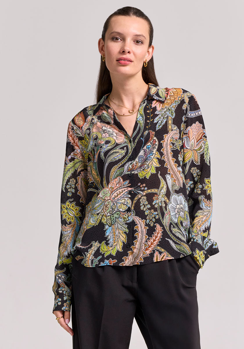 A woman with straight brown hair wears a Princess Goes Hollywood Paisley-Print Hemdbluse in green, orange, blue, and yellow on black. She stands facing forward with one hand in the pocket of her black pants.