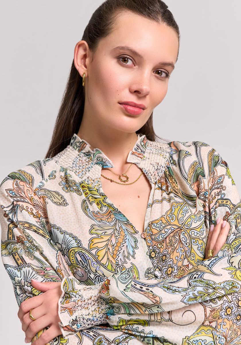 A woman with light skin and straight brown hair pulled back poses with arms crossed, wearing a Princess Goes Hollywood paisley-print blouse with a smocked stand collar, gold hoop earrings, and layered necklaces against a light background.