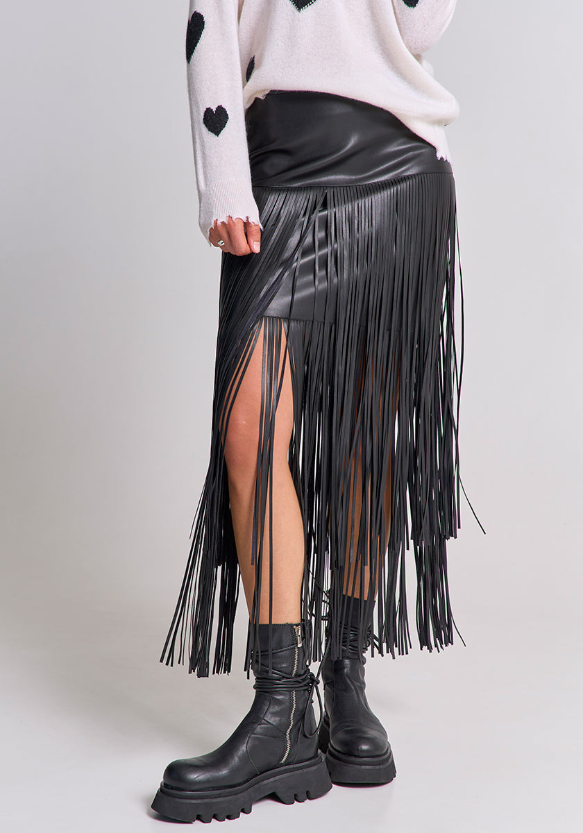 A person wears a white sweater with black heart patterns, a Princess Goes Hollywood black faux leather mini skirt with long fringes and back zipper, and black chunky combat boots. The photo focuses on the lower body against a plain background.