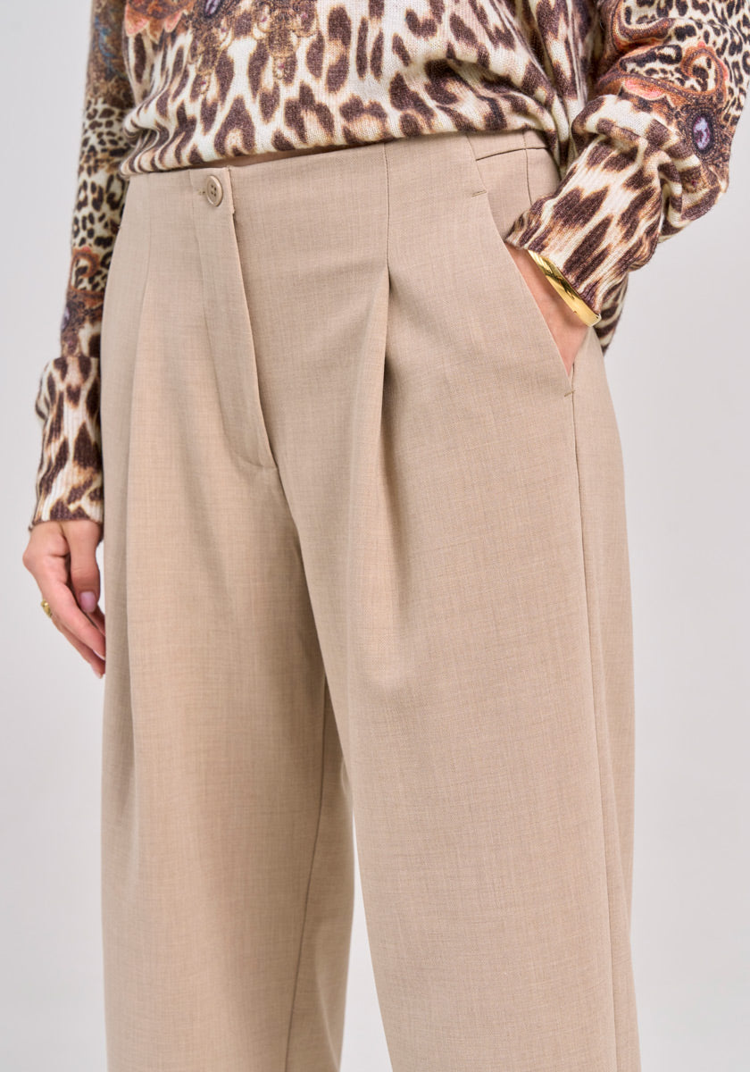 A person stands with one hand in the pocket of beige 7/8-length wide-leg trousers by Princess Goes Hollywood, featuring front pleats. They wear a tucked-in leopard print top, gold bracelet, and ring against a plain background.