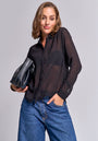 Eine Frau mit kurzen braunen Haaren lächelt in einer langen, transparenten Hemdbluse von Princess Goes Hollywood über einem schwarzen BH, kombiniert mit blauen Jeans. Sie hält eine schwarze Clutch und posiert vor grauem Hintergrund.