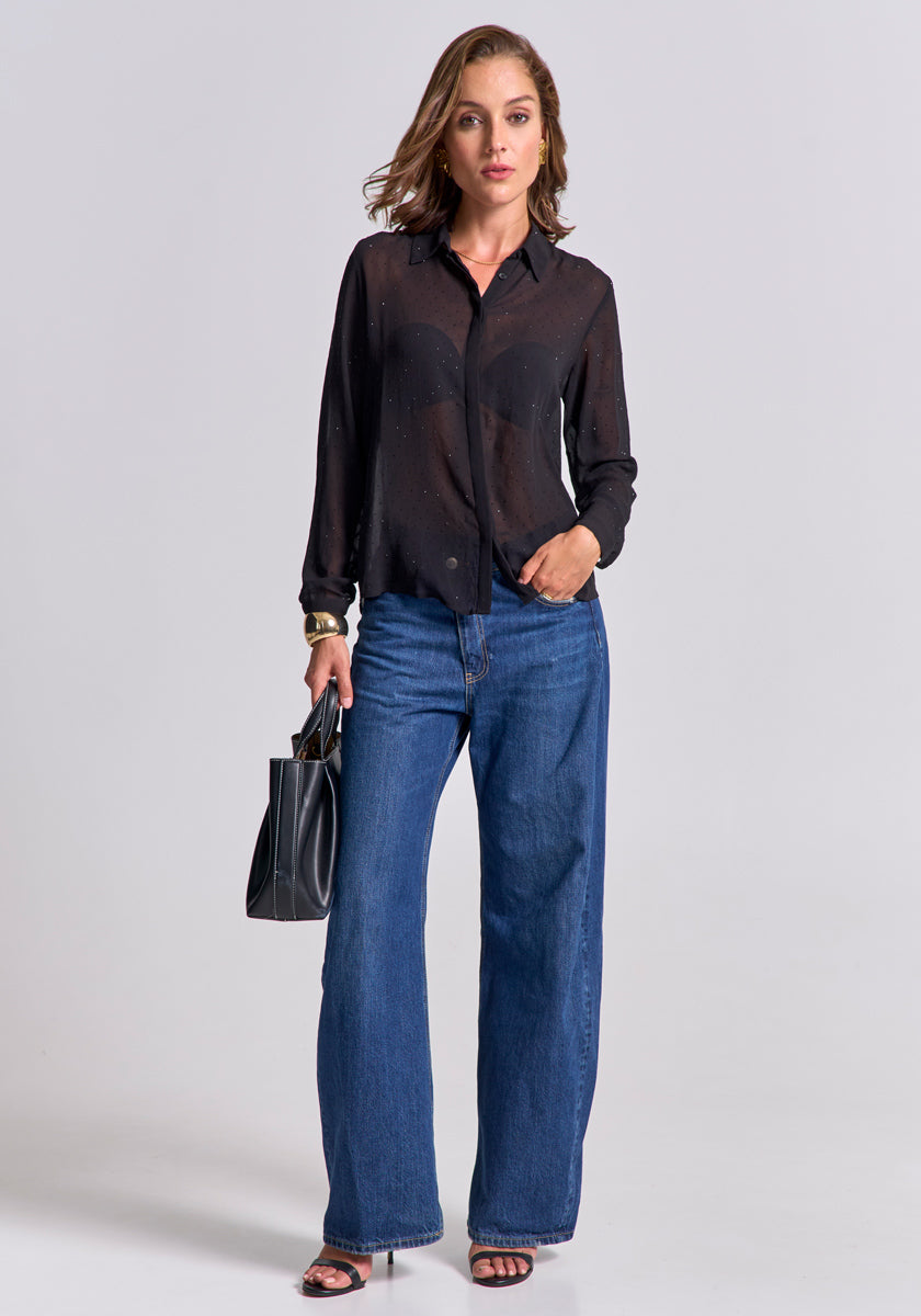 A woman with loose waves poses against a light gray background in a Princess Goes Hollywood Transparente Hemdbluse mit langen Ärmeln, wide-leg blue jeans, and black heeled sandals. She accessorizes with a black handbag and a large gold bracelet.