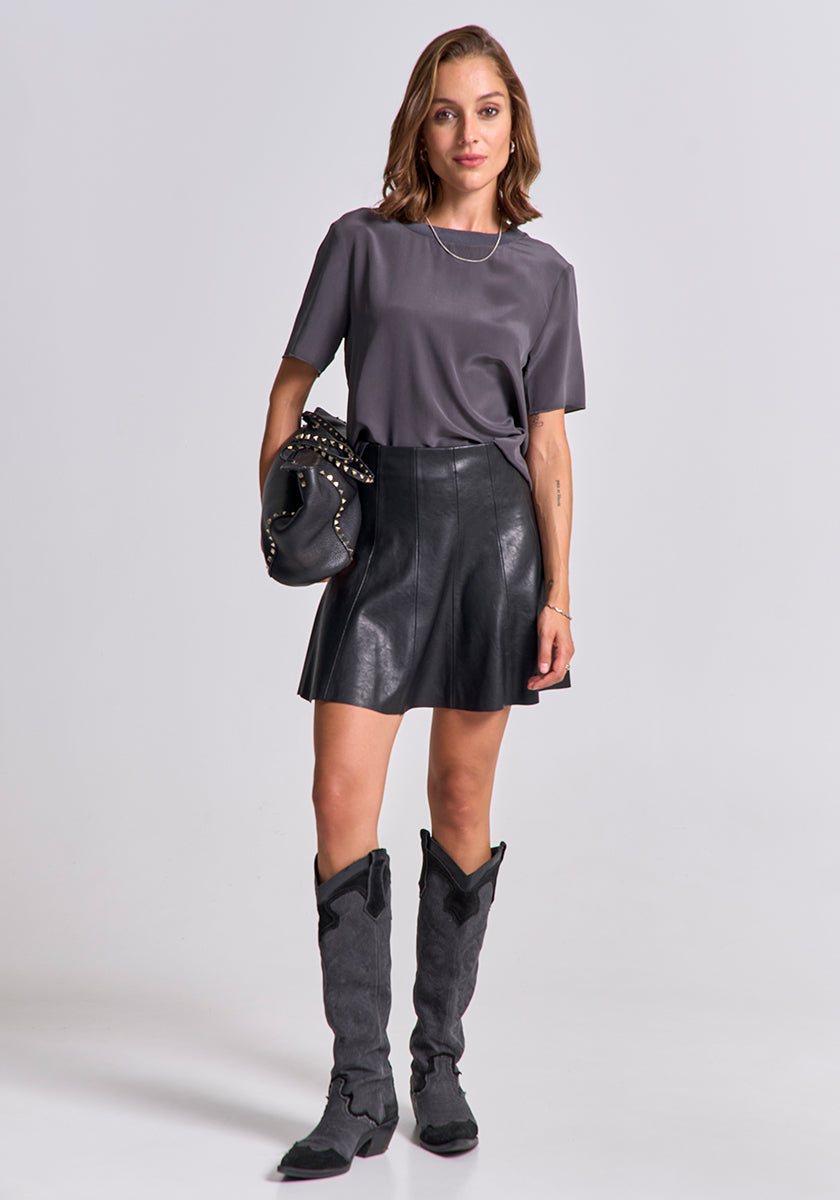 A woman stands against a plain light gray background, wearing a graues Princess Goes Hollywood Damen T-Shirt mit elastischem Rundhalsausschnitt, einem schwarzen Leder-Minirock in A-Linie und hohen Cowboy-Boots. Sie hält eine schwarze Clutch und hat wellige, schulterlange Haare.