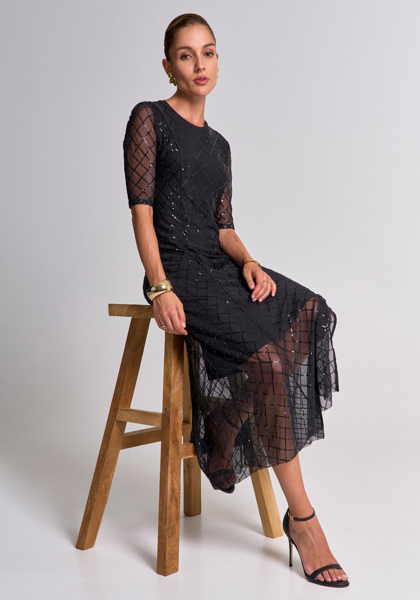 Eine Frau sitzt auf einem Holzstuhl vor schlichtem Hintergrund und trägt ein schwarzes Meshkleid mit Pailletten im geometrischen Muster von Princess Goes Hollywood. Goldene Ohrringe, Armband und High Heels vervollständigen den eleganten Look.