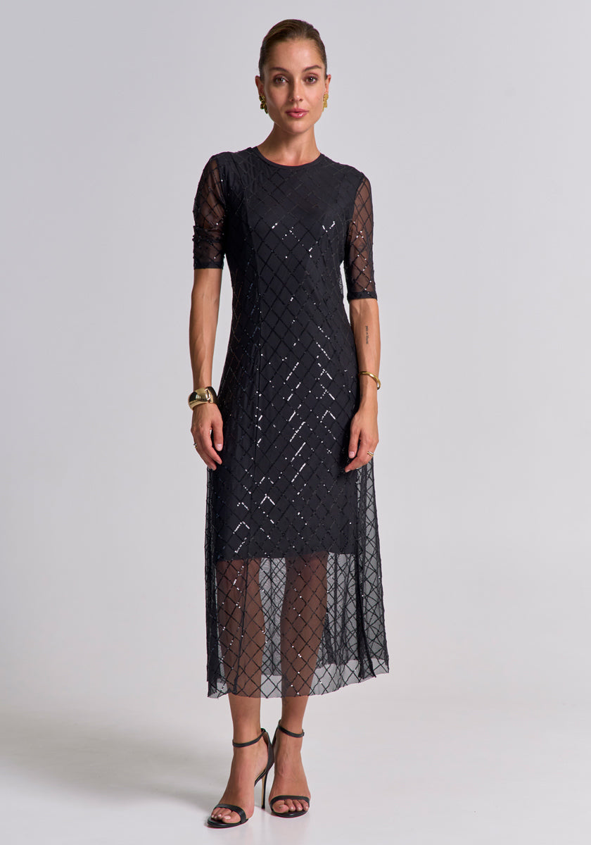 A woman stands facing forward against a plain background, wearing a calf-length, diamond-patterned schwarzes Meshkleid by Princess Goes Hollywood with short sleeves. She pairs it with black high-heeled sandals and gold jewelry, her hair pulled back.