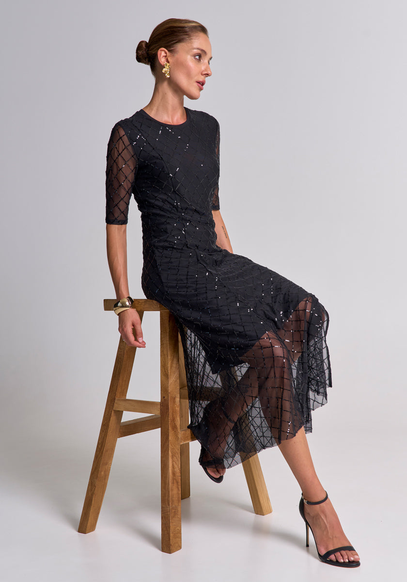 A woman with brown hair in a sleek bun sits on a wooden stool, wearing a schwarzes Midilanges Meshkleid mit Pailletten von Princess Goes Hollywood, schwarze Riemchenheels, goldene Ohrringe und ein Armband vor schlicht-grauem Hintergrund.