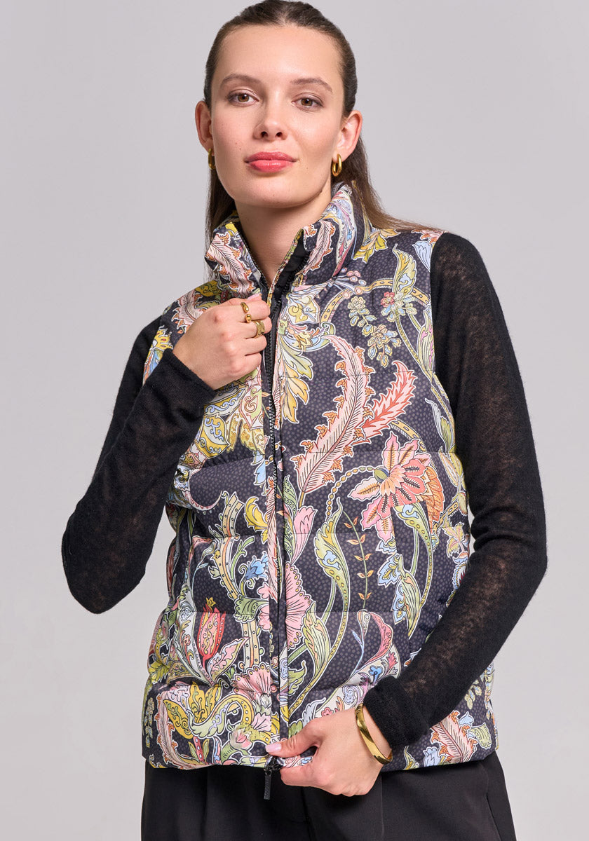 A woman with light skin and straight brown hair, pulled back, wears the Princess Goes Hollywood Paisley-Print Steppweste mit Zwei-Wege-Reissverschluss over a black long-sleeve top. She softly smiles at the camera, holding the vest collar against a plain light gray background.