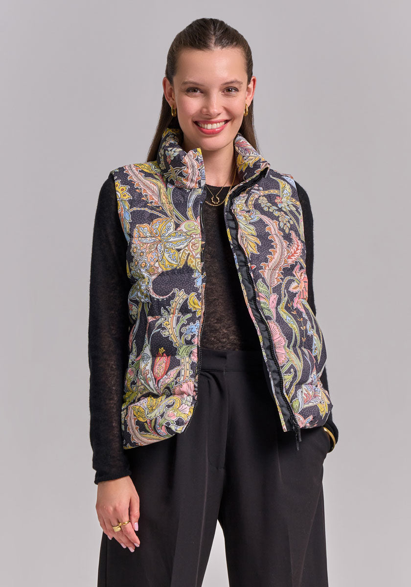 A woman with straight brown hair smiles, wearing a Princess Goes Hollywood Paisley-Print Steppweste mit Stehkragen over a black long-sleeve shirt and high-waisted pants, standing against a plain light gray background with her hands in her pockets.
