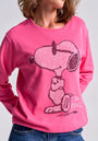 A woman models a Princess Goes Hollywood Snoopy sweatshirt with rhinestone embellishments and "too cool" in white below the cartoon dog. She pairs it with blue jeans, standing with her hands behind her back.