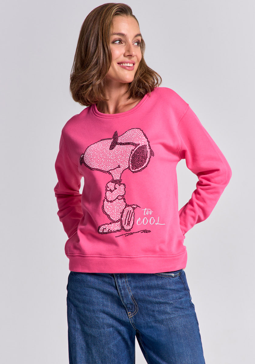 A woman with shoulder-length brown hair smiles to the side, wearing a bright pink Princess Goes Hollywood Snoopy-print sweatshirt with &quot;too cool&quot; text and blue jeans, standing against a plain light gray background.