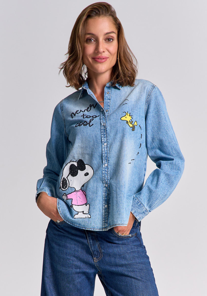 A woman with shoulder-length brown hair smiles at the camera, hands in jean pockets, wearing a light blue Princess Goes Hollywood cotton shirt featuring Snoopy in sunglasses, a pink shirt, Woodstock flying, and the phrase "Never too cool.