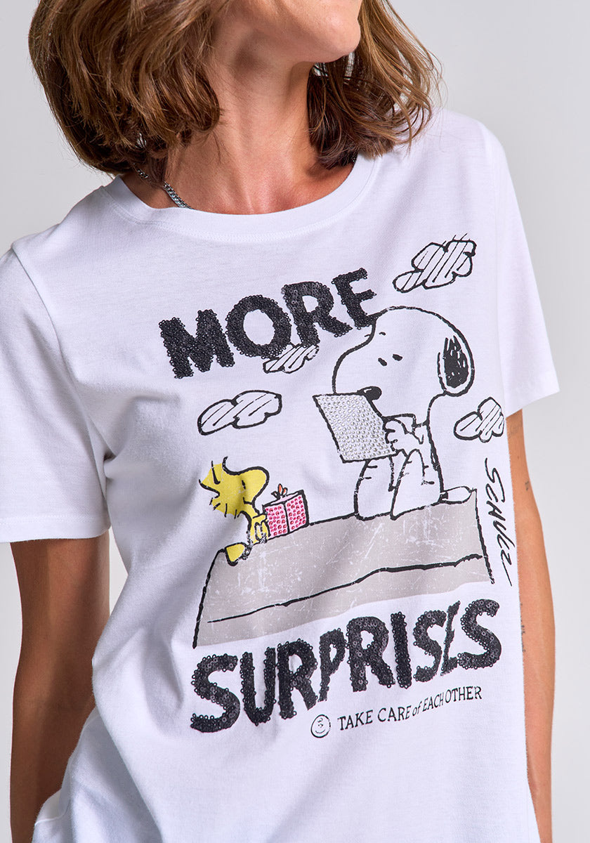 A woman wears a Princess Goes Hollywood Peanuts-print T-shirt, with Snoopy reading a letter, Woodstock holding a gift, clouds, Peanuts creator’s signature, and text: “MORE SURPRISES” and “TAKE CARE OF EACH OTHER.”.