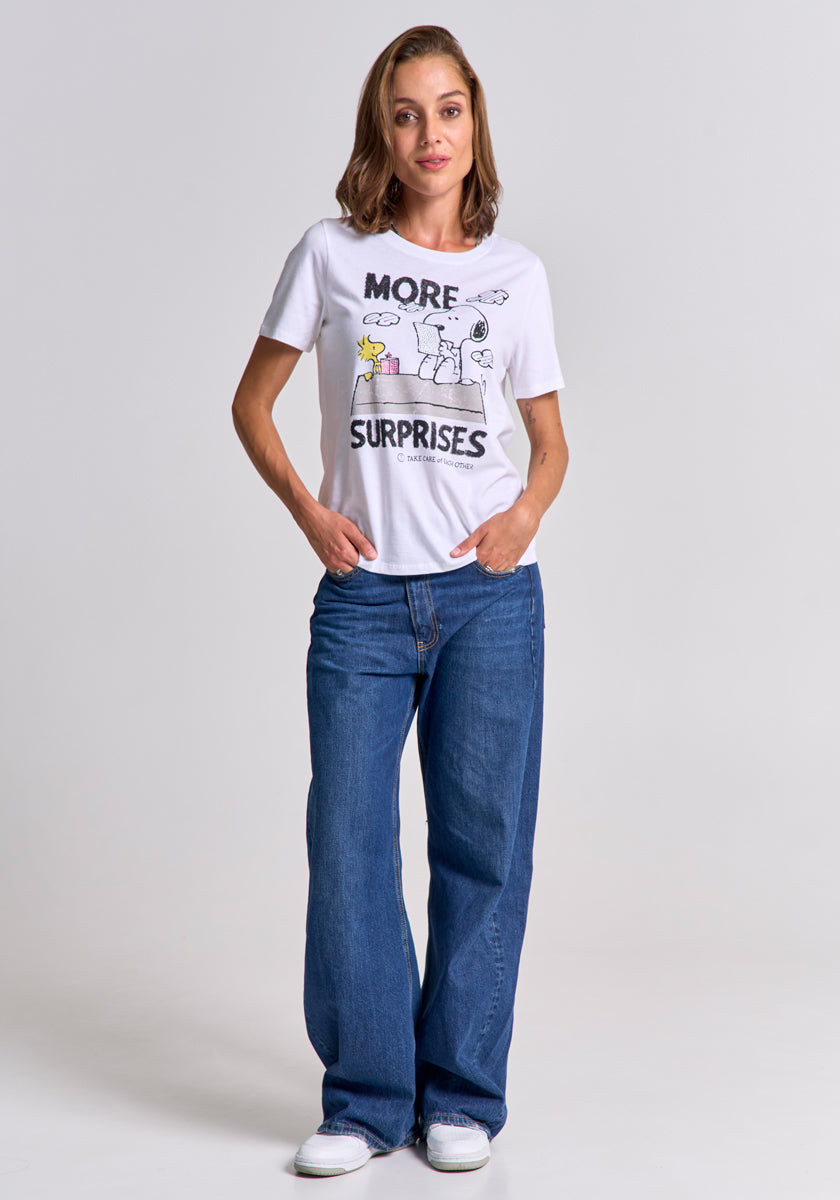 A woman with light brown hair stands facing forward, smiling slightly, wearing a Princess Goes Hollywood white Peanuts-print T-shirt with MORE SURPRISES and cartoon graphics, blue wide-leg jeans, and white sneakers. Her hands are in her pockets.