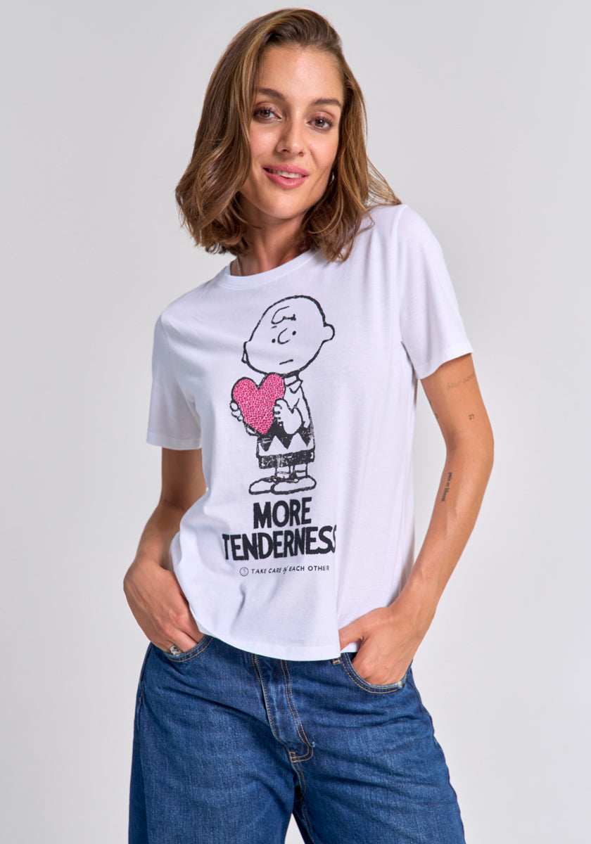 A woman with shoulder-length brown hair wears a Princess Goes Hollywood Charlie Brown cotton T-shirt, showing him holding a pink heart and "MORE TENDERNESS" text. She smiles gently with one hand in her blue jeans pocket against a plain light background.