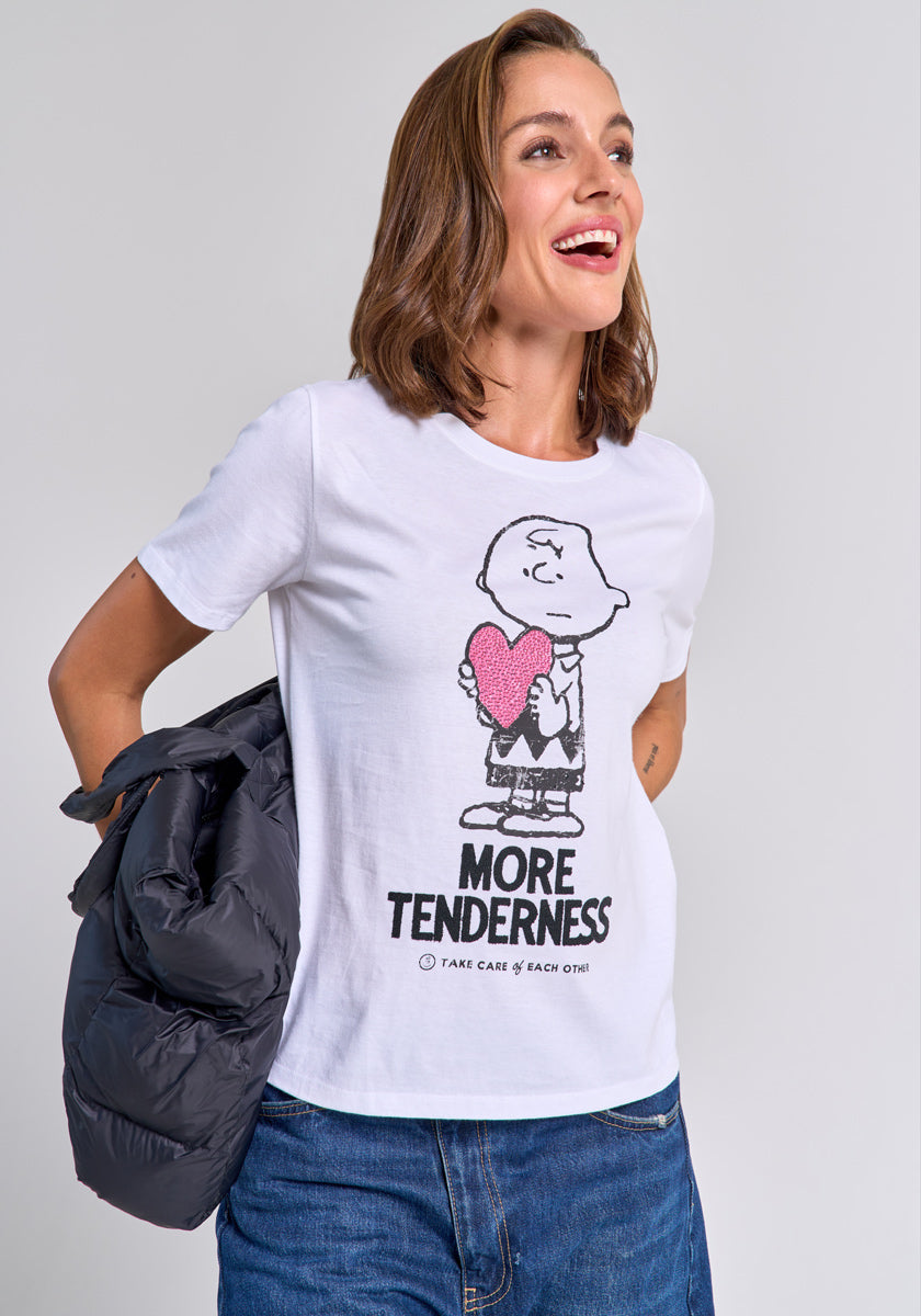 A smiling woman with light brown hair wears a Princess Goes Hollywood Charlie Brown T-shirt featuring a cartoon character holding a pink heart and the text "MORE TENDERNESS." She holds a black jacket, wears blue jeans, and stands against a gray background.