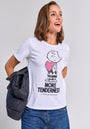 A smiling woman with light brown hair wears a Princess Goes Hollywood Charlie Brown T-shirt featuring a cartoon character holding a pink heart and the text "MORE TENDERNESS." She holds a black jacket, wears blue jeans, and stands against a gray background.