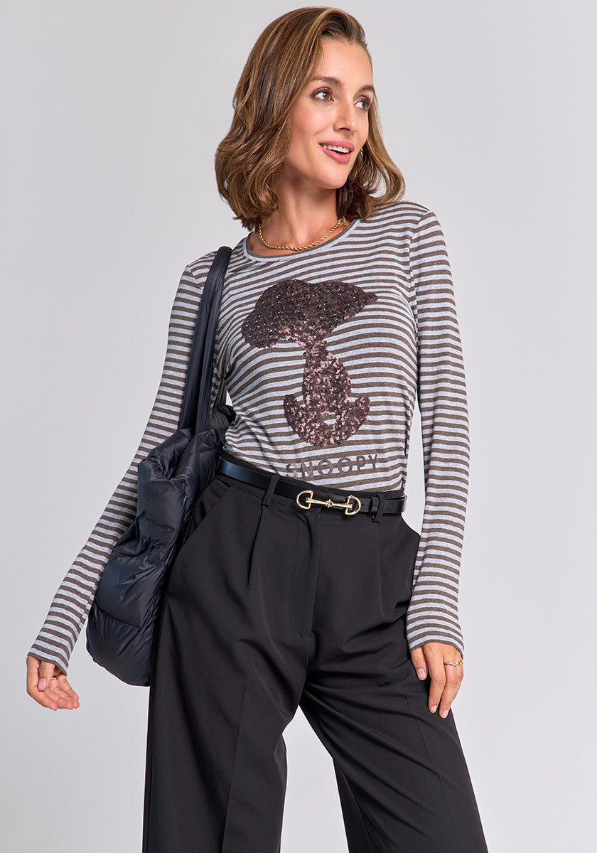 A woman with wavy, shoulder-length hair wears a Princess Goes Hollywood striped long-sleeve shirt with Snoopy motif and sequin details, tucked into belted high-waisted black trousers. She carries a black padded bag on her shoulder and smiles, looking to the side.