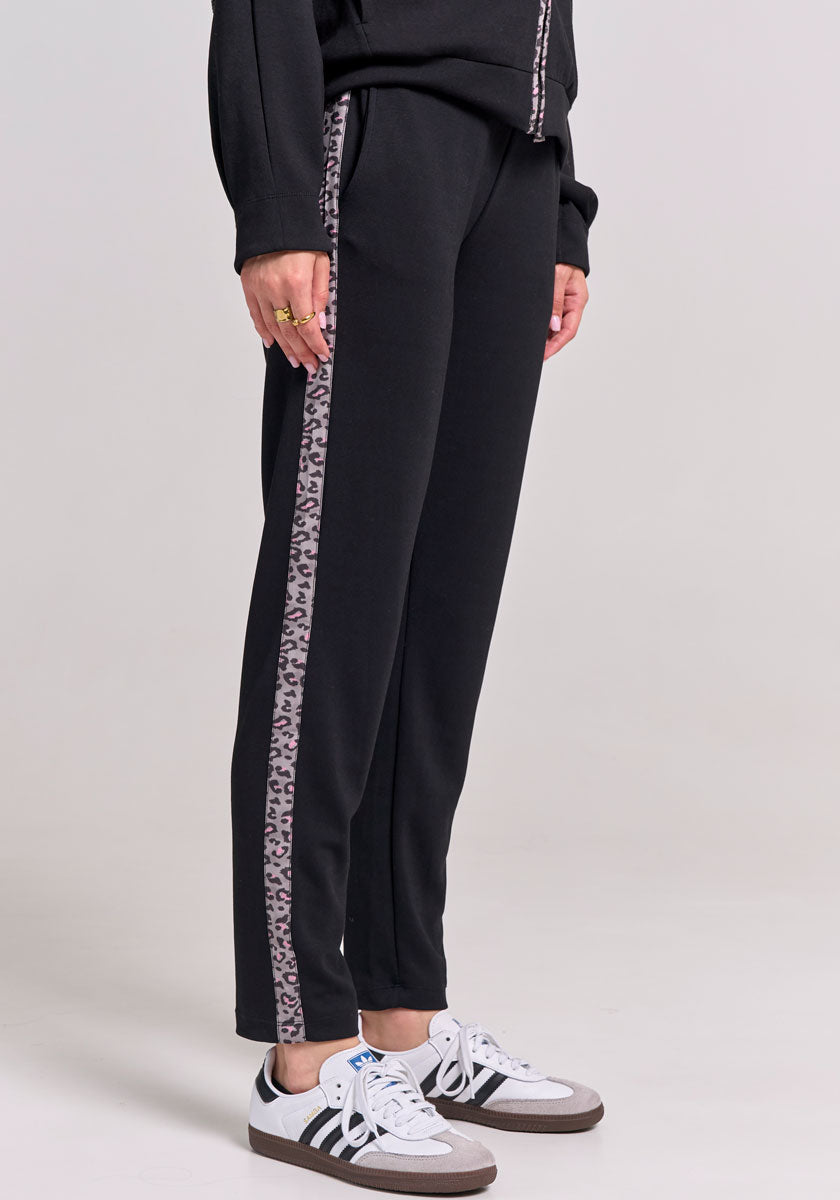 A person wears Frogbox by Princess Modalmix Sweathose with a vertical leopard stripe, paired with a black top and white Adidas sneakers with black stripes and gray accents, shown from the waist down on a light gray background.